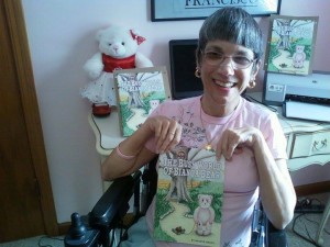 (Image description: Joanne, a woman with gray hair and glasses wearing a pink shirt sitting in a wheelchair holding a book she wrote entitled, “The Busy World of Bianca Bear” which has a cartoon bear on the cover)