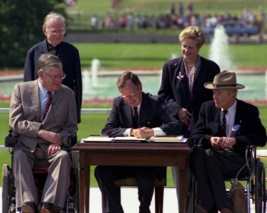 (Image Description: Photo of President George H. W. Bush signing into law the Americans with Disabilities Act of 1990 on the South Lawn of the White House. Evan Kemp and Justin Dart are in wheelchairs beside President Bush. Rev. Harold Wilke and Swift Parrino are standing behind President Bush.)