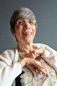 [Image description: Joanne, a woman with short gray hair and wire frame glasses smiling at the camera. She is wearing a light colored top with a floral print. Her hands are leaning up against her chest.]